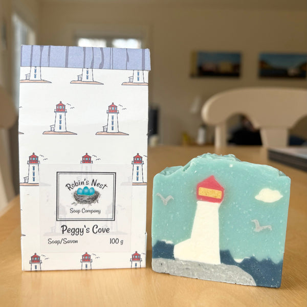 A handmade soap bar shaped like a lighthouse scene sits on a wooden table next to its branded paper packaging. The soap is light blue with a white lighthouse, dark blue waves, and a red roof.